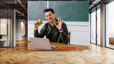 smiling male teacher in glasses holding apple and showing ok sign at computer desk in classroom Wall mural