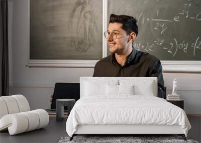 smiling male teacher sitting at desk and showing laptop with blank screen in classroom Wall mural