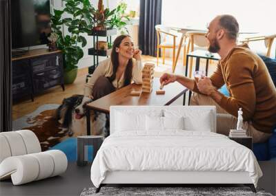 Smiling man holding coffee and playing wood blocks game with girlfriend near border collie at home Wall mural