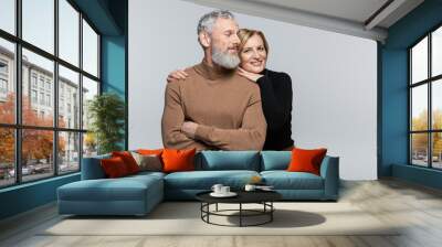 Smiling mature woman in turtleneck hugging husband and looking at camera isolated on grey. Wall mural