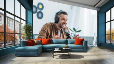 smiling radio host in headphones gesturing while speaking in microphone in broadcasting studio Wall mural