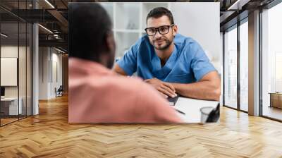 smiling rehabilitologist in eyeglasses listening to blurred african american patient Wall mural