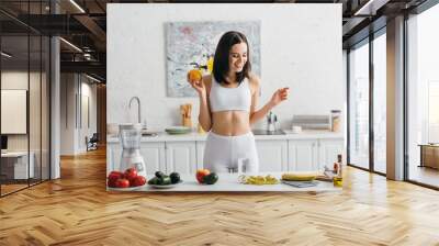 Smiling sportswoman weighing fruits near glass of water and measuring tape on kitchen table, calorie counting diet Wall mural