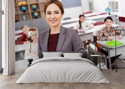 Smiling teacher with digital tablet standing in front of pupils and looking at camera Wall mural