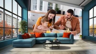 Smiling woman feeding pasta to son near husband and orange juice in kitchen Wall mural