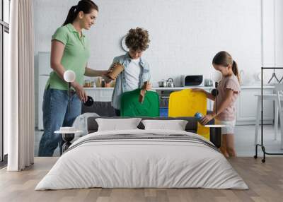 Smiling woman standing near kids with garbage and trash cans with recycle emblem at home Wall mural