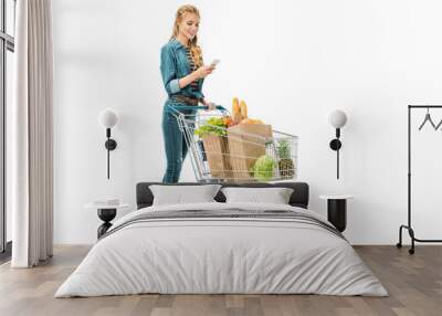 smiling woman using smartphone and carrying shopping trolley with products isolated on white Wall mural