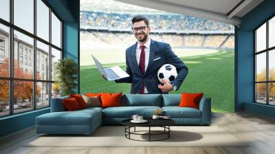 smiling young businessman in suit with laptop and soccer ball sitting on football pitch at stadium, sports betting concept Wall mural