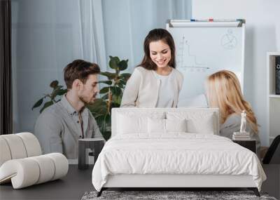 smiling young businesswoman looking at male and female colleagues sitting with papers at desk in office Wall mural