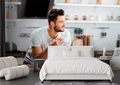 smiling young man holding cup of coffee and leaning at kitchen table at morning Wall mural