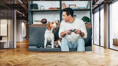 Smiling young man with gamepad sitting on sofa and looking at dog Wall mural