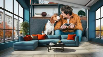 Smiling young man with gamepad sitting on sofa and looking at dog Wall mural