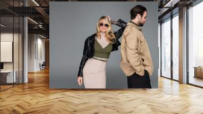 stylish blonde woman in sunglasses and leather jacket leaning on back of bearded man isolated on grey Wall mural