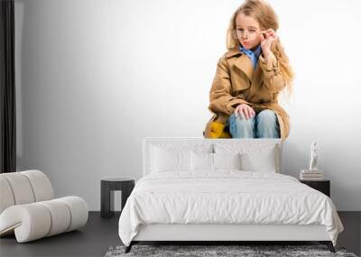 Stylish kid in glasses wearing trench coat and sitting on suitcase isolated on white Wall mural