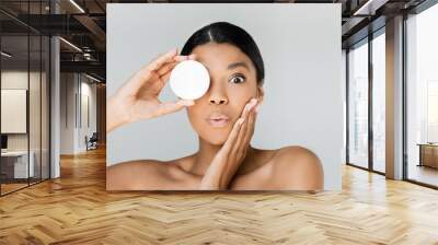 surprised african american woman covering eye while holding container with face cream isolated on grey Wall mural