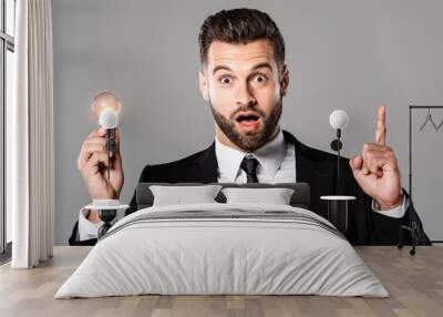 surprised businessman in black suit holding glowing light bulb and showing idea gesture isolated on grey Wall mural