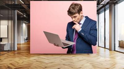 thoughtful businessman in suit using laptop and holding hand near face on pink background Wall mural