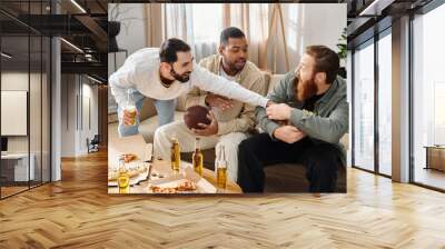 Three cheerful, interracial friends in casual attire sit around a table, laughing and sharing pizza and beer. Wall mural