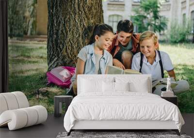 three cute classmates reading book and smiling while sitting on lawn under tree Wall mural