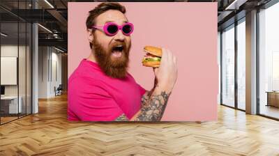 thrilled man in trendy sunglasses opening mouth near tasty burger isolated on pink. Wall mural