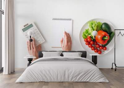 Top view of woman using calculator near notebook and fresh vegetables on plate on white background Wall mural