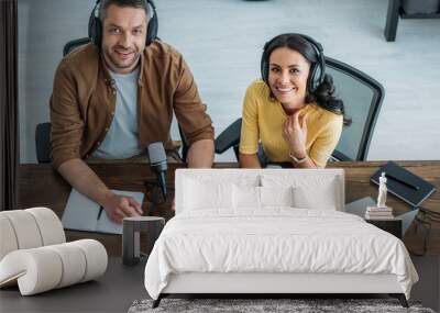 two cheerful radio hosts in headphones sitting at workplace and smiling at camera Wall mural