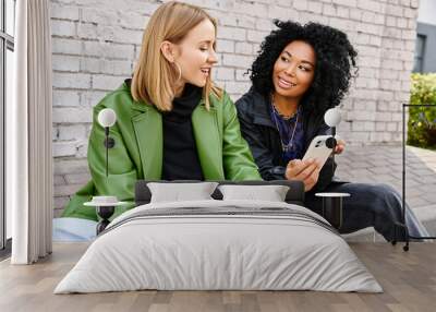 Two diverse women in casual attire sit on a bench, engrossed in a cell phone. Wall mural