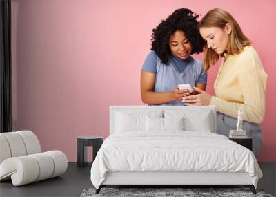 Two diverse women in casual attire standing together, engrossed in a cell phone. Wall mural