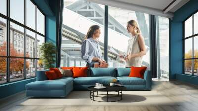 two female coworkers smiling and looking at each other, holding phone and working papers, coworking Wall mural