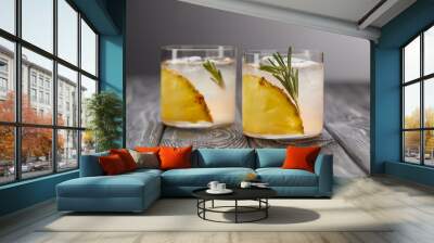 two glasses of lemonade with pineapple pieces, ice cubes and rosemary on grey wooden tabletop Wall mural