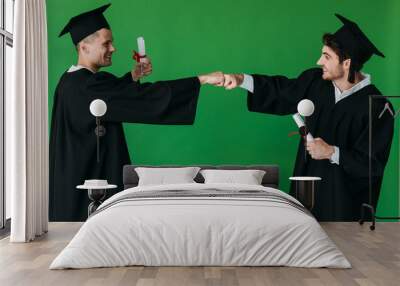 two happy students in academic caps holding diplomas and touching fists isolated on green Wall mural