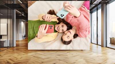 Two pretty teenage girls in casual attire, laying on a bed, taking selfies with their cell phones. Wall mural