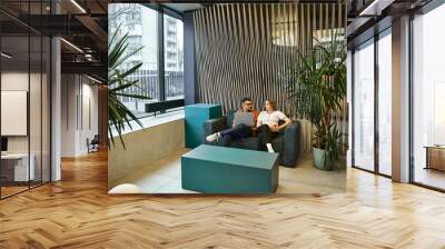 Two professionals from a startup team sit on a trendy couch in a contemporary office space, taking a break from work. Wall mural