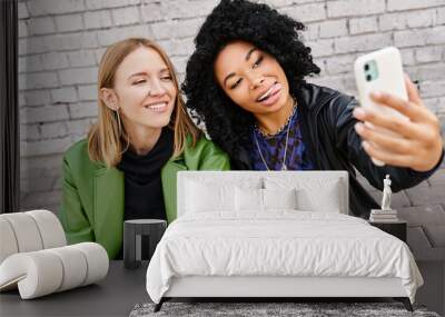 Two women in casual attire sitting on the ground, capturing a moment with a selfie. Wall mural