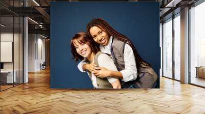 Two young women, hugging joyfully in stylish attire against a dark blue background. Wall mural