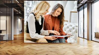 Two young women, one bride-to-be, engrossed in book while sitting on couch, sharing a moment of connection. Wall mural