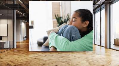 upset african american teenage girl hugging knees while sitting with closed eyes at home Wall mural