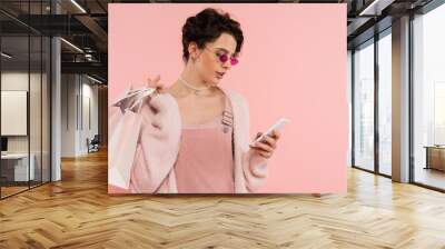 woman in trendy sunglasses holding shopping bags and messaging on smartphone isolated on pink Wall mural