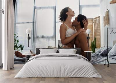 Young, sexy couple of diverse backgrounds showing love and affection by hugging and kissing passionately on a bed. Wall mural