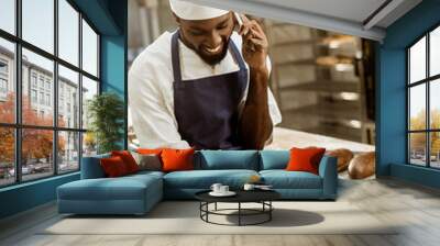 young african american baker with hands covered in flour talking by phone while sitting on work table Wall mural