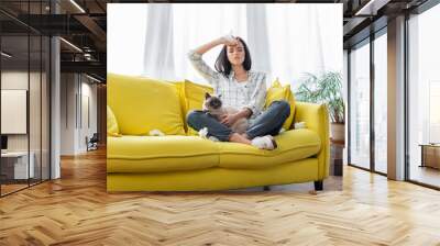 young allergic woman sitting on sofa with cat and suffering from headache Wall mural