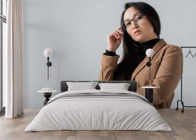 young asian woman in beige blazer looking at camera while adjusting glasses isolated on grey Wall mural