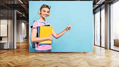 young blonde student with backpack holding notebooks and ponging with finger on blue Wall mural