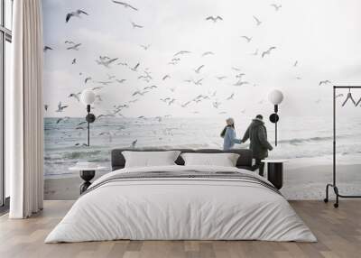 young couple holding hands and walking on winter sea shore and looking at seagulls Wall mural