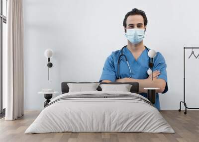 young doctor in medical mask standing with crossed arms and looking at camera isolated on grey. Wall mural