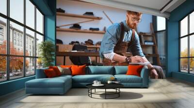 young male fashion designer in apron and eyeglasses taking notes at workshop Wall mural