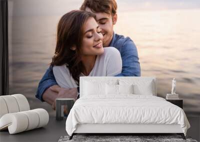 Young man hugging brunette girlfriend with closed eyes beside sea at sunset Wall mural