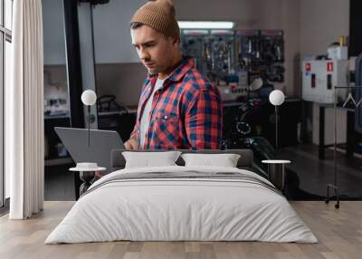 young mechanic in plaid shirt and beanie using laptop in workshop near motorcycle on blurred background Wall mural