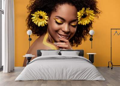 Young sensual african american woman with artistic make-up and gerberas in hair isolated on orange background Wall mural