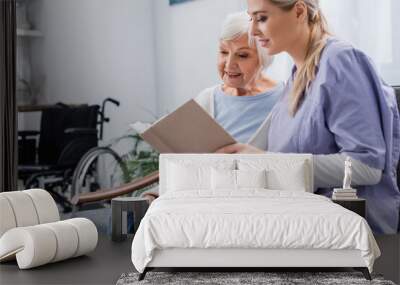 young social worker reading book to senior woman while sitting on sofa Wall mural